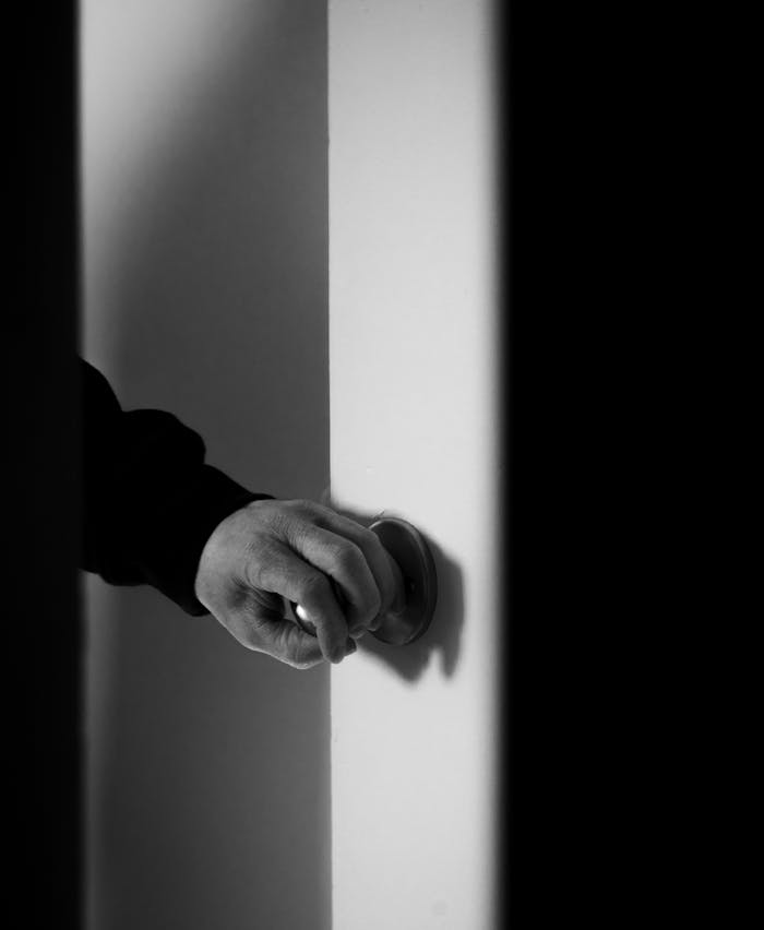A black and white close-up of a hand opening a door, evoking mystery and curiosity.